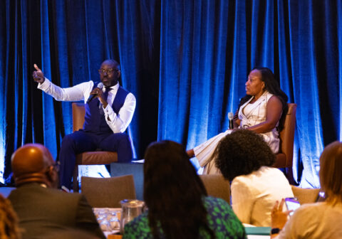 Man in vest and white shirt speaks with microphone next to woman in white outfit.
