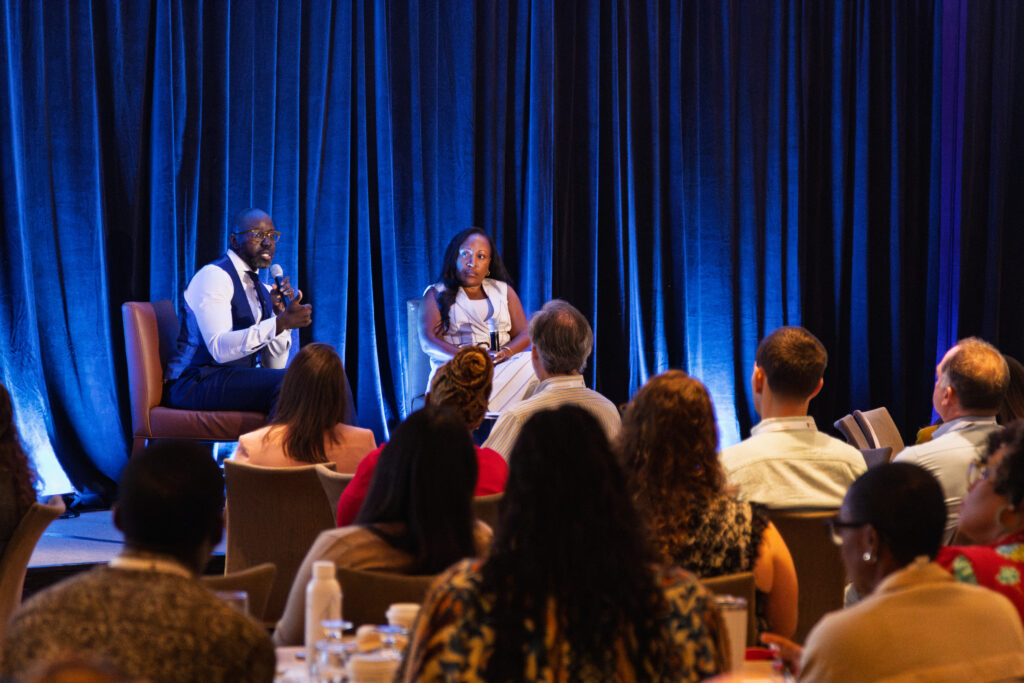 HCZ CEO seated on stage with a colleague,, speaking in front of a crowd of attentive conference attendees.