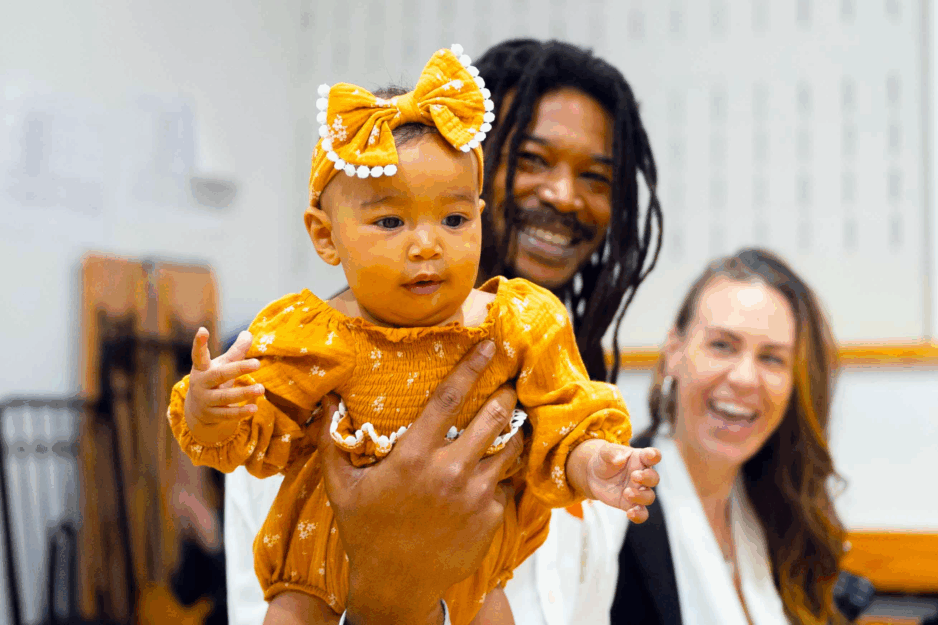 Man and woman smiling as the man holds a baby girl with one hand towards the camera.