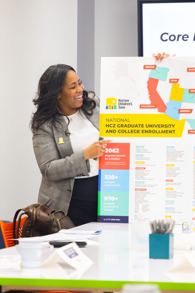 A woman dressed in business professional attire smiles as she presents at a map at the front of the room