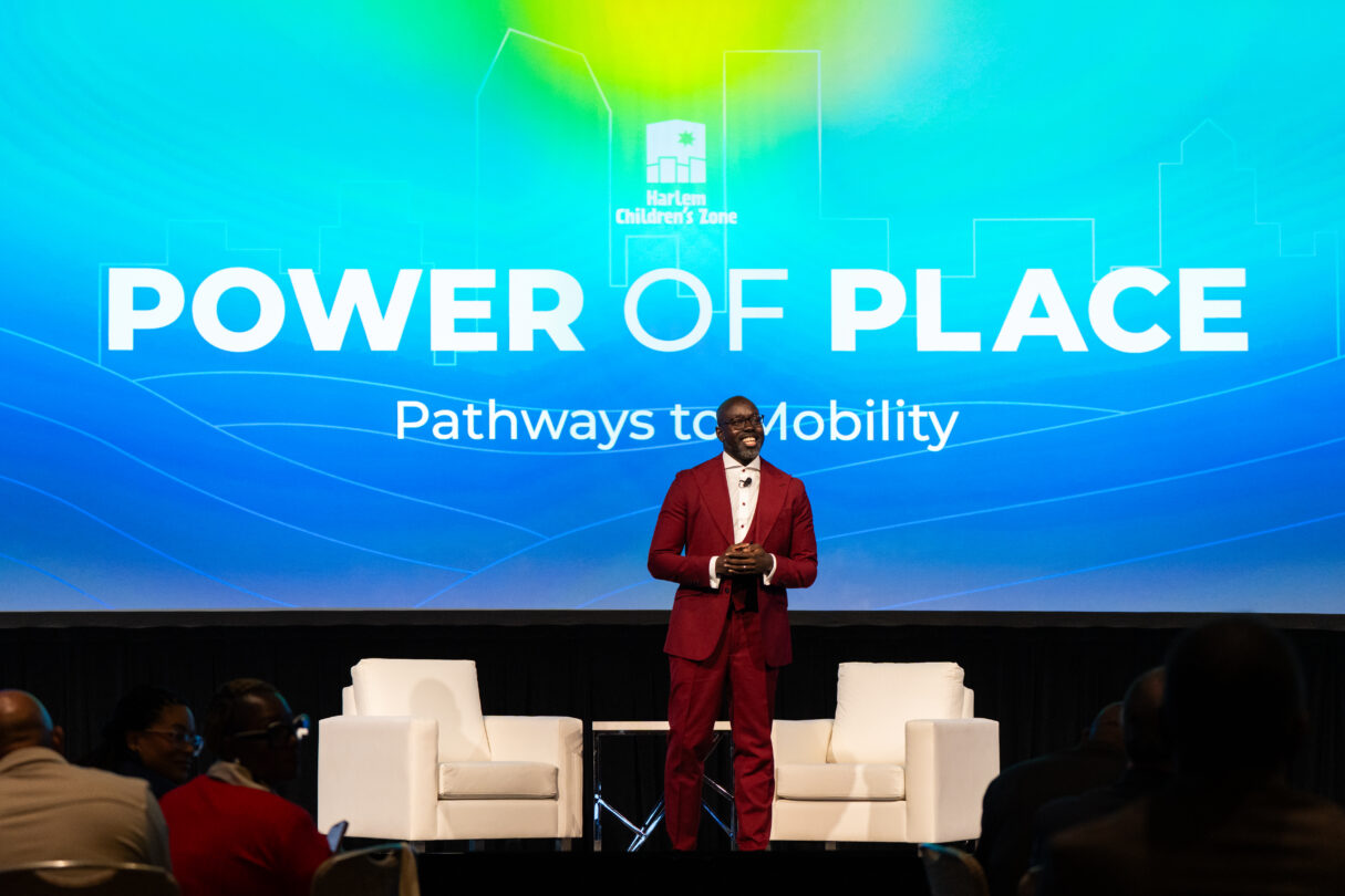 Man in maroon suit standing on stage in front of a screen with the words "Power of Place" projected on it.
