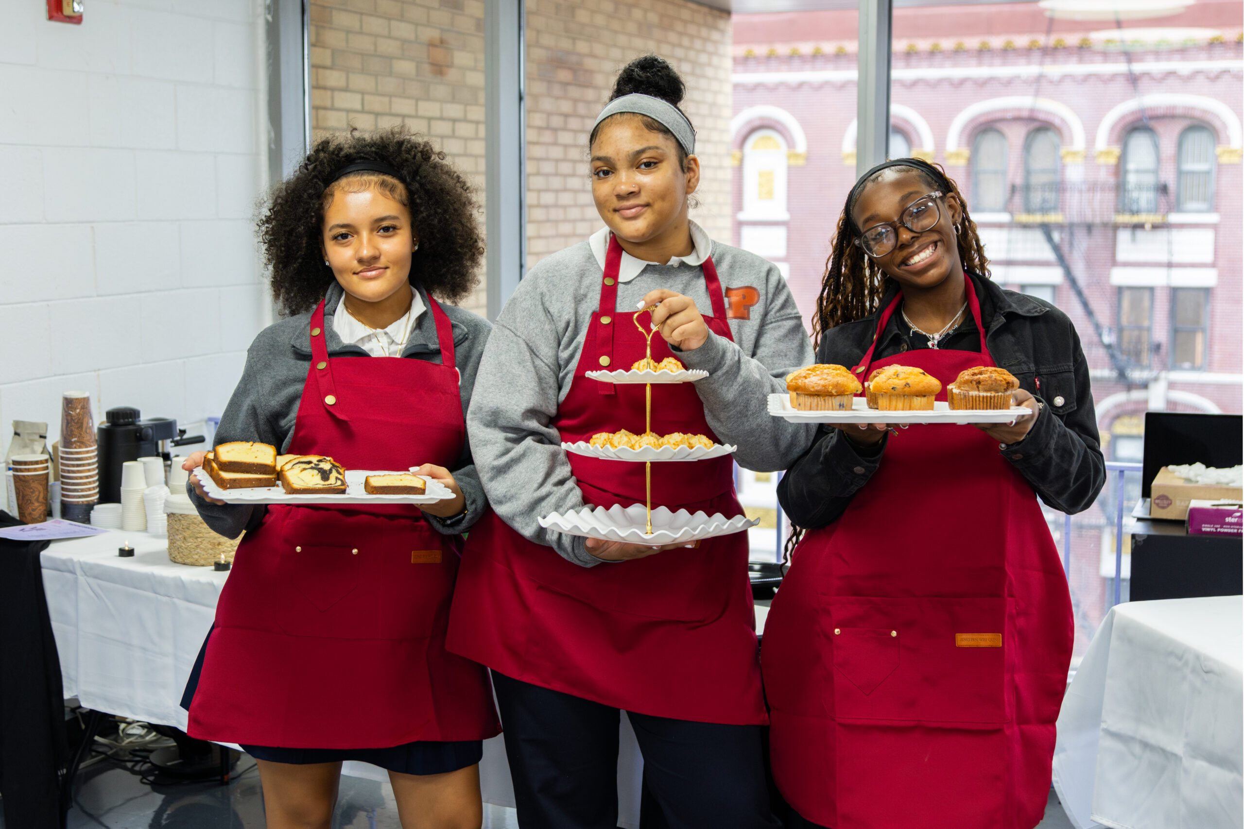 Three young women wearing matching red aprons smile as the hold trays of food.
