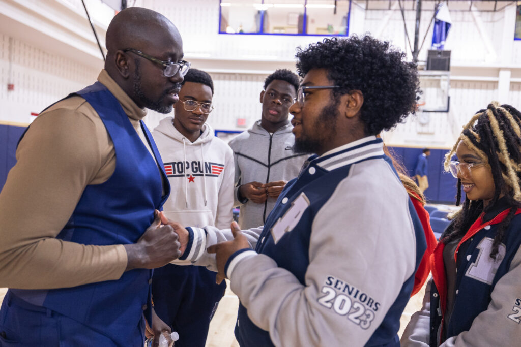 Man in blue vest shakes hand of man in blue and grey jacket in gymnasium.