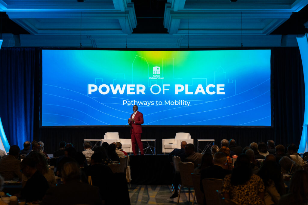 Man in maroon suit stands on stage in hotel conference room and speaks to attendees.