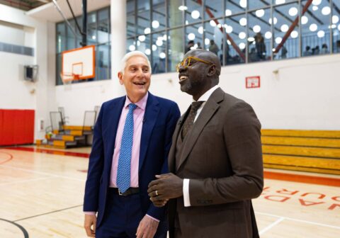 Man in brown suit and man in navy blue suit talk in gymnasium.