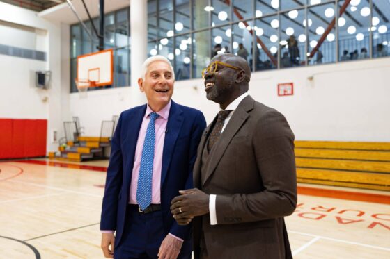 Man in brown suit and man in navy blue suit talk in gymnasium.
