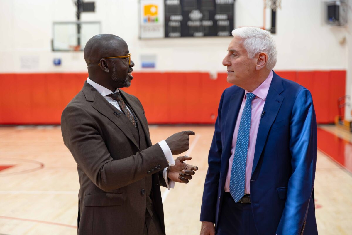 Man in brown suit talks to man in navy blue suit in gymnasium.