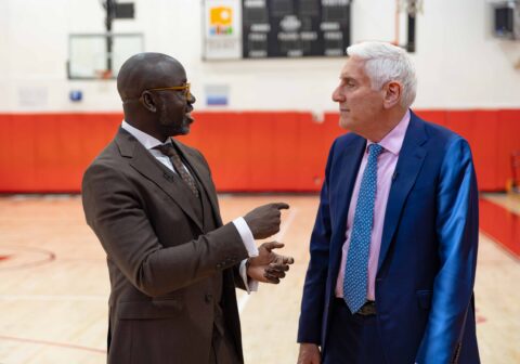 Man in brown suit talks to man in navy blue suit in gymnasium.