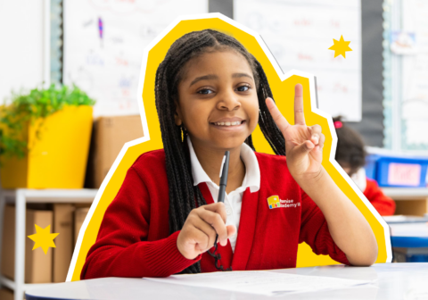 Young female scholar smiling and holding up a peace sign in a classroom setting.