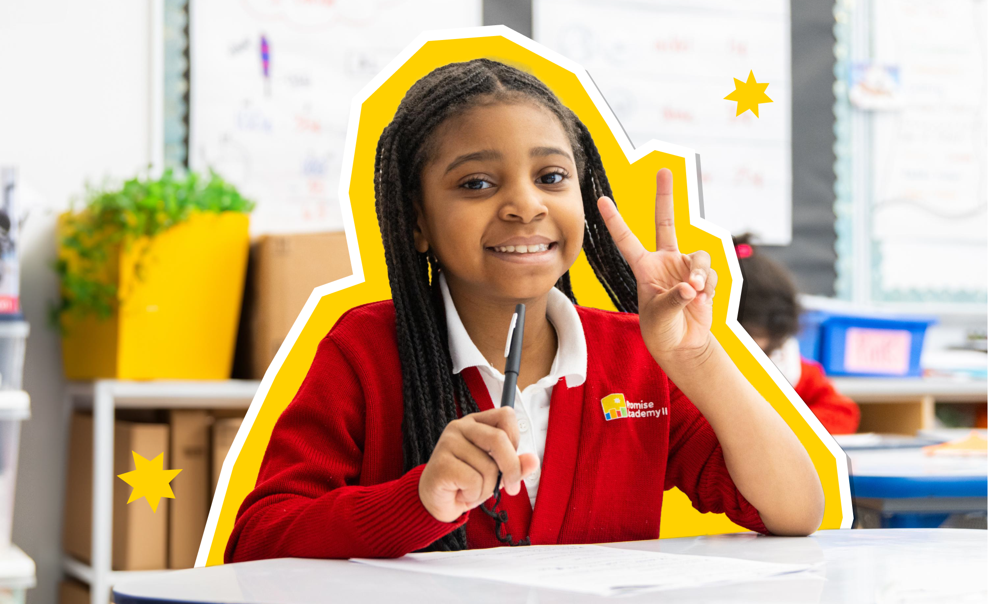 Young female scholar smiling and holding up a peace sign in a classroom setting.